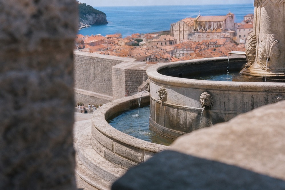 A wide stone fountain with water flowing from carved mask spouts into a large pool, with old city walls behind it