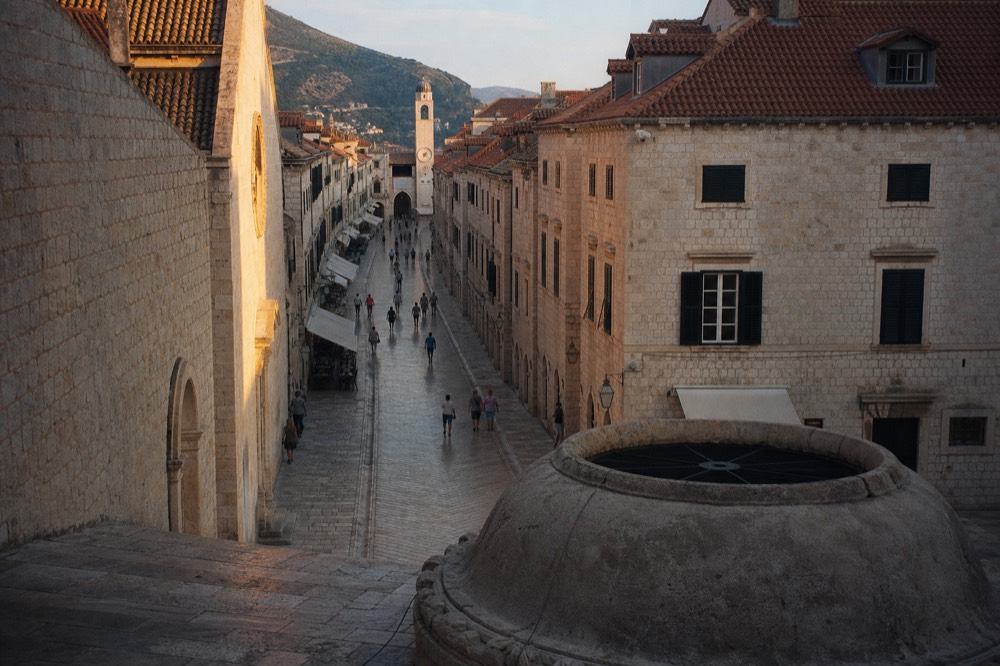 The long limestone main street of a walled old town seen from a stone fountain toward a distant clock tower