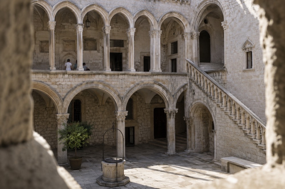 Stone courtyard of a Gothic palace with an arched colonnade and a staircase leading to a mezzanine