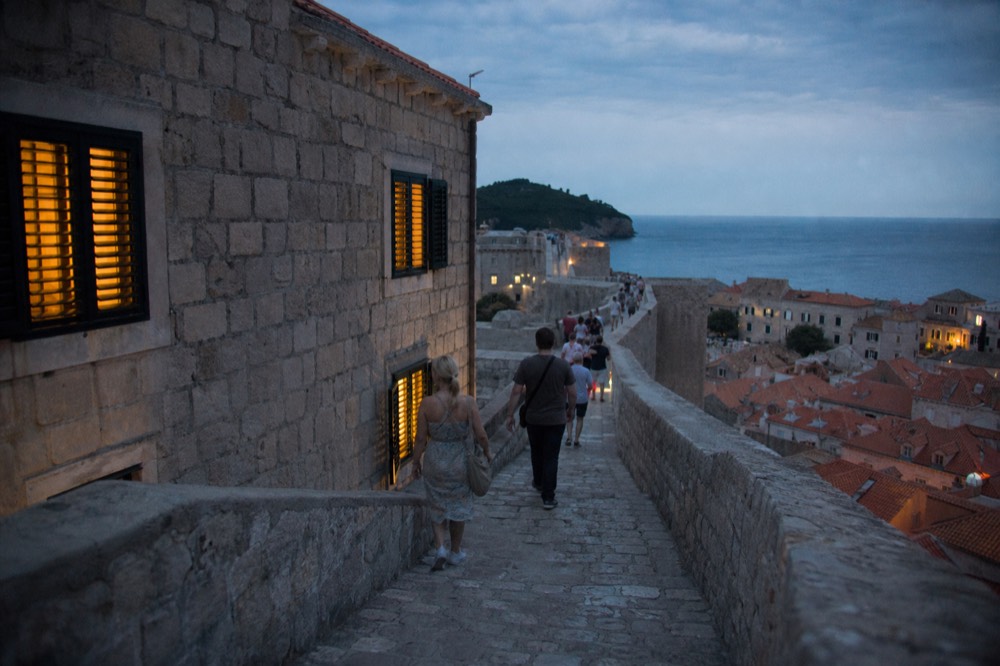 Visitors walking along the top of the old city walls above red rooftops and the sea