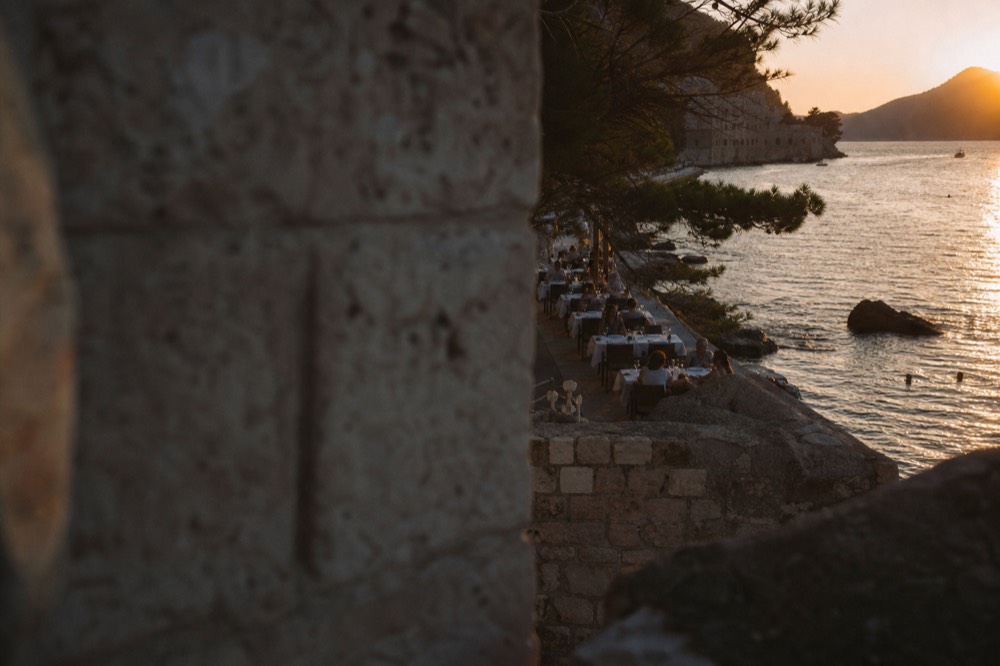 Waterfront restaurant tables along a pine-fringed bay promenade at dusk