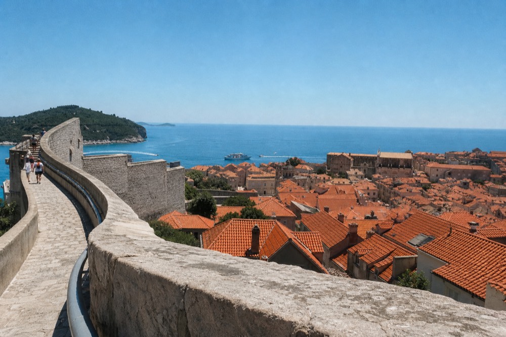The stone city walls of Dubrovnik curving above red rooftops with the Adriatic Sea beyond