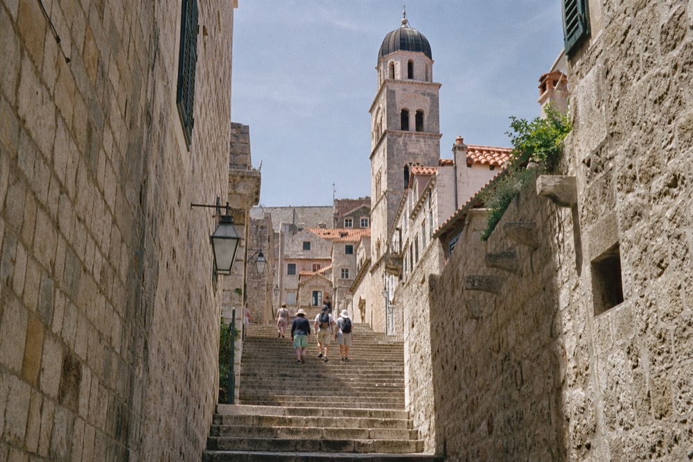 A steep stone staircase street in Dubrovnik Old Town rising toward a tower in bright sunlight