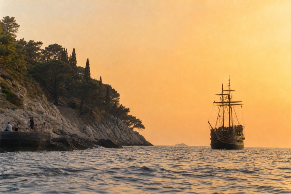 A wooden sailing ship with masts silhouetted against an orange sky above the sea