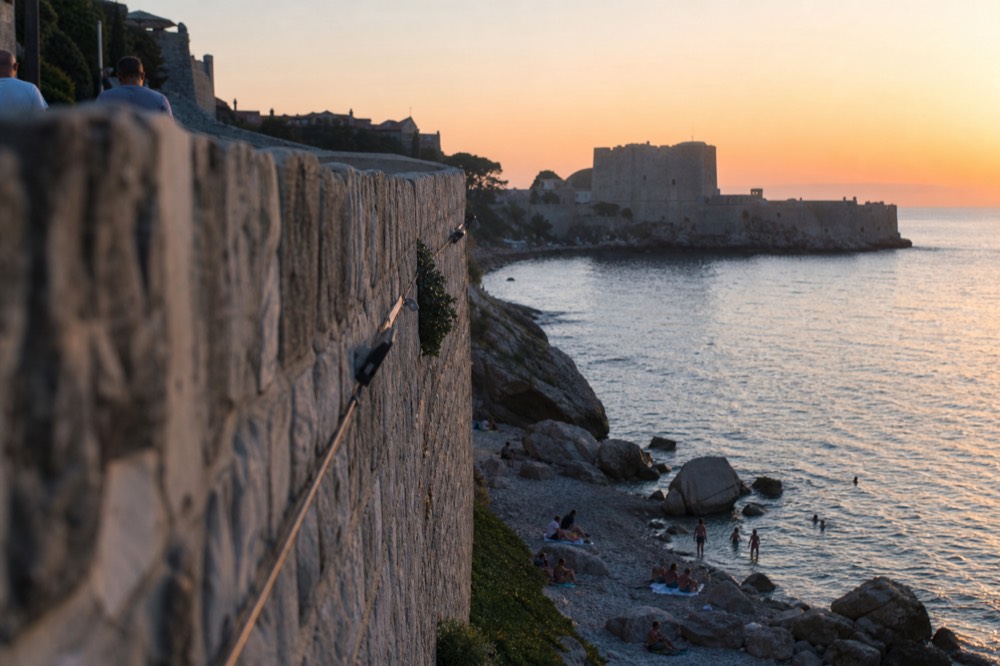 A stone terrace above the sea with a view of a fortress silhouetted against an orange evening sky