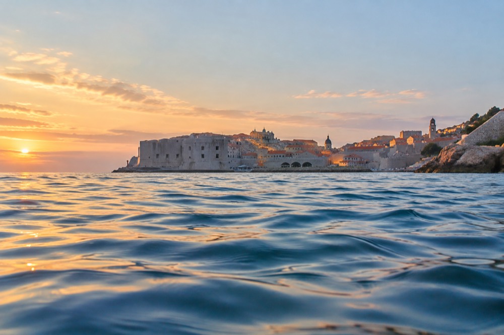 Swimmer's view across calm blue water toward a walled city at golden hour