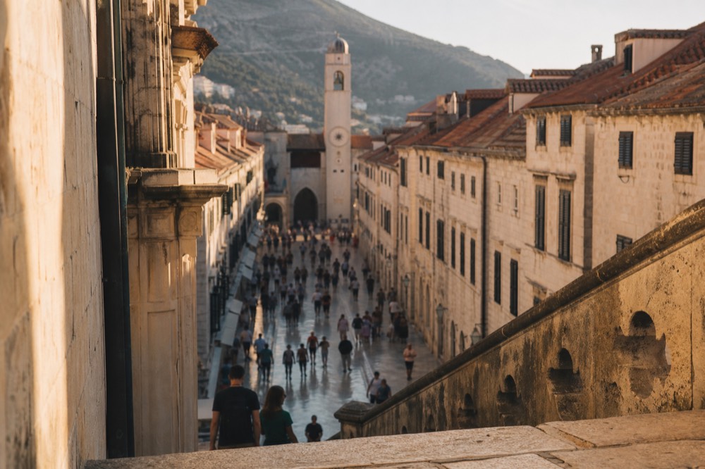 Crowds of tourists walking along the Stradun on a busy afternoon, with the clock tower visible.