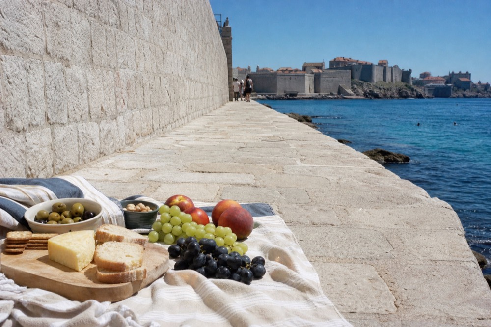 A vegetarian picnic spread on a beach towel with bread, cheese, olives, and fresh fruit, overlooking the Adriatic.