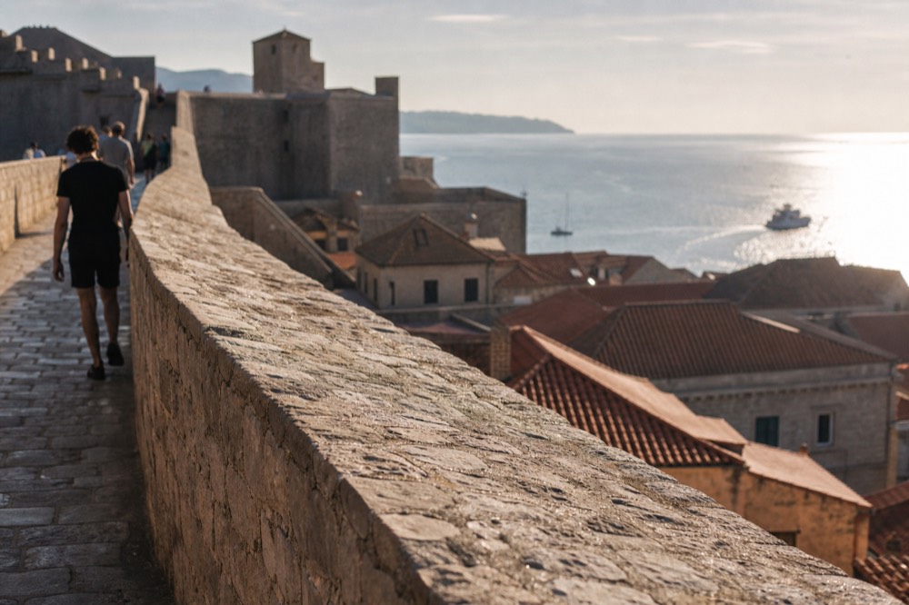 Walkers on a stone city wall path above red roofs with the sea beyond