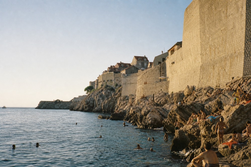 Swimmers in clear turquoise water at a rocky beach below Dubrovnik's walls