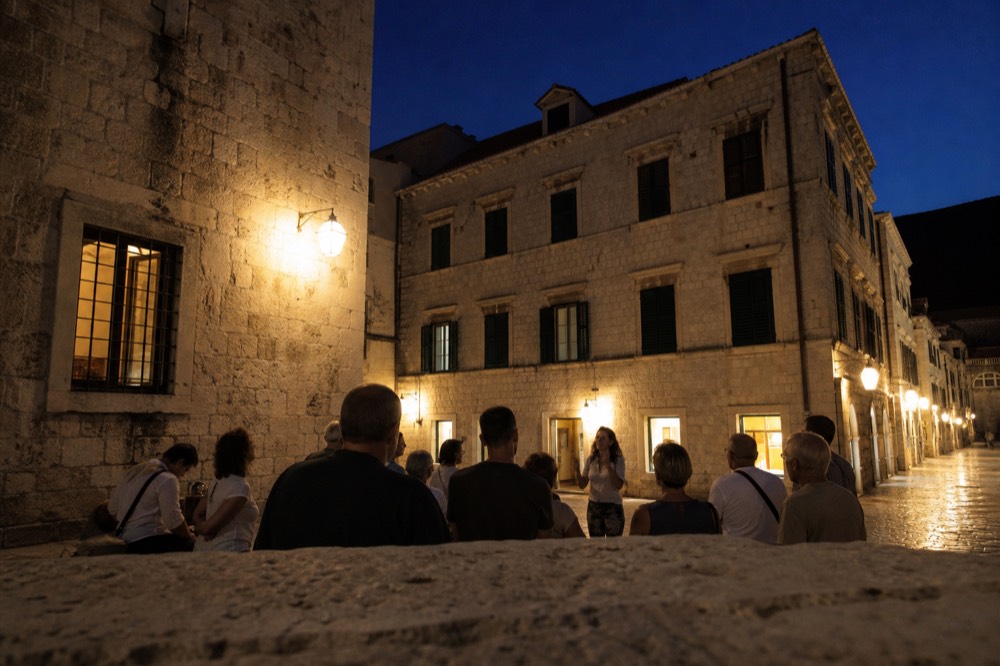 A small group listening to a guide in a lamplit Dubrovnik square at night