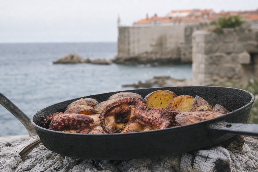 An octopus and potato peka lifted from a cast iron pan over embers