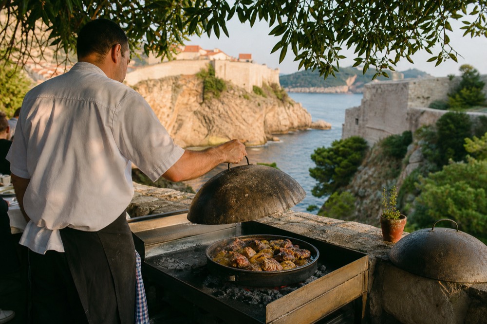A cook lifting the domed steel lid off a peka pan over glowing embers