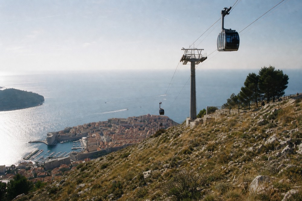 Cable car cabins above a hillside with the walled city and sea below