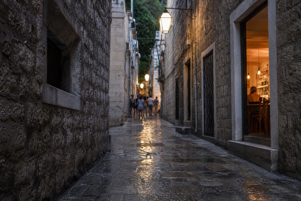 A narrow stone alley in Dubrovnik Old Town at dusk with warm light from bar windows